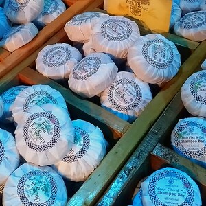May include: A close-up of a wooden crate filled with round, white, and blue shampoo bars. The bars are wrapped in paper with a blue and white pattern and have the text "Shampoo Bar" printed on them.