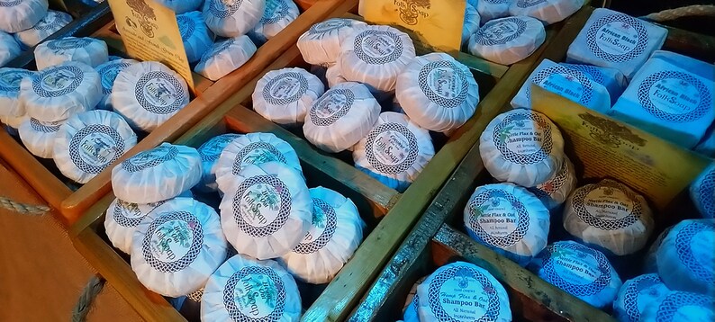 May include: A close-up of a wooden crate filled with round, white, and blue shampoo bars. The bars are wrapped in paper with a blue and white pattern and have the text "Shampoo Bar" printed on them.