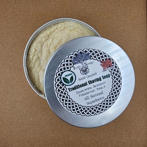 Op de afbeelding: Een zilveren blik met een wit etiket waarop staat "Folk Soap Hand Crafted Traditional Shaving Soap Sandalwood Bergamot Frankincense Vanilla All Natural Ingredients" met een groen vegan-logo en een rood drakenlogo. Het blik is open en toont een witte scheerschuim erbinnen.
