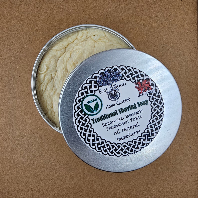 Op de afbeelding: Een zilveren blik met een wit etiket waarop staat "Folk Soap Hand Crafted Traditional Shaving Soap Sandalwood Bergamot Frankincense Vanilla All Natural Ingredients" met een groen vegan-logo en een rood drakenlogo. Het blik is open en toont een witte scheerschuim erbinnen.