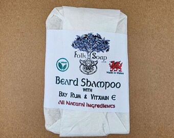 Op de afbeelding: Een witte zeepstaaf gewikkeld in wit papier. Het etiket vermeldt "Folk Soap" met een groen vegan-logo, een rood drakenlogo en de tekst "Made in Wales". De zeep is gelabeld met "Beard Shampoo with Bay Rum & Vitamin E. All Natural Ingredients."