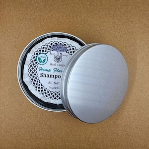 May include: A silver tin with a white label that reads "Folk Soap Hand Crafted Hemp Flax Shampoo All Natural Ingredients Vegan". The tin is open and the lid is sitting next to it.
