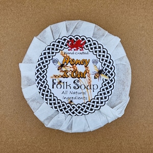 May include: A round bar of white soap wrapped in white paper. The label features a black and white Celtic knot design with a red dragon, a wheat stalk, and two bees. The text on the label reads "Hand Crafted Honey & Oat Folk Soap All Natural Ingredients".