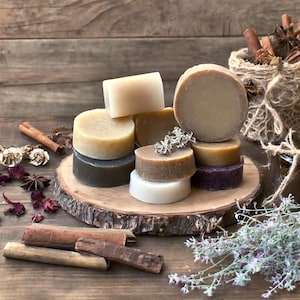 May include: A collection of round, natural soap bars in various shades of brown, beige, and white, arranged on a wooden slice. The soap bars are surrounded by cinnamon sticks, dried flowers, and a burlap bag.
