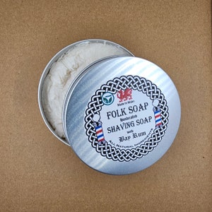 May include: A silver tin of Folk Soap handcrafted shaving soap with Bay Rum scent. The tin has a Celtic knot design and a red dragon logo. The label reads "Folk Soap Handcrafted Shaving Soap with Bay Rum All Natural Ingredients Made in Wales Vegan".