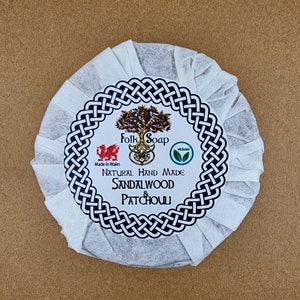 May include: A white round bar of soap wrapped in white tissue paper. The soap has a black and white Celtic knot design around a white label with the text "Folk Soap", a red dragon logo, the text "Made in Wales", a green leaf logo, the text "Vegan", and the text "Natural Hand Made Sandalwood & Patchouli".