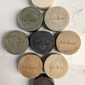 May include: Eight round bars of soap with the words "Folk Soap" stamped on each bar. The bars are arranged in a circle with the white bar at the top and the brown bar at the bottom. The bars are on a white marble surface.