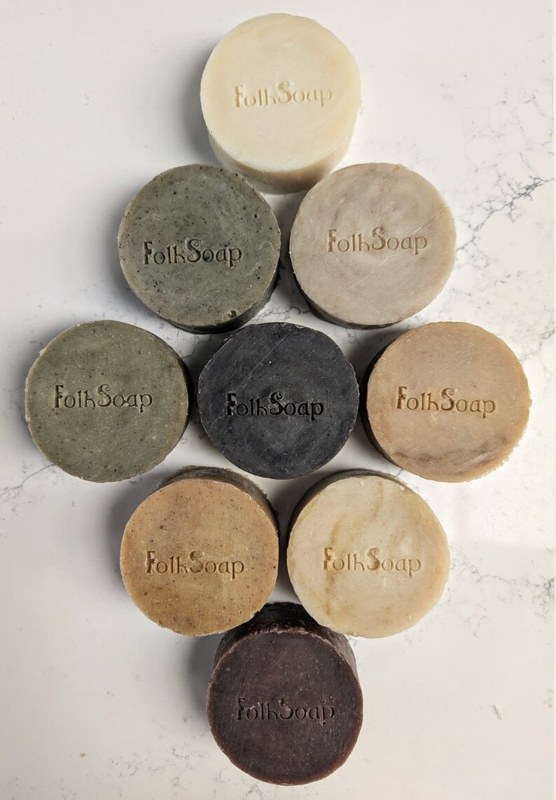 May include: Eight round bars of soap with the words "Folk Soap" stamped on each bar. The bars are arranged in a circle with the white bar at the top and the brown bar at the bottom. The bars are on a white marble surface.