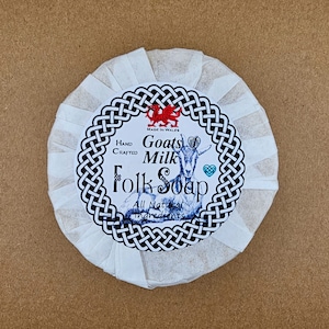 May include: A white round bar of goat milk soap with a black and white label that says "Hand Crafted Goats Milk Folk Soap" with a red dragon and a goat illustration. The label also says "Made in Wales" and "All Natural Ingredients".