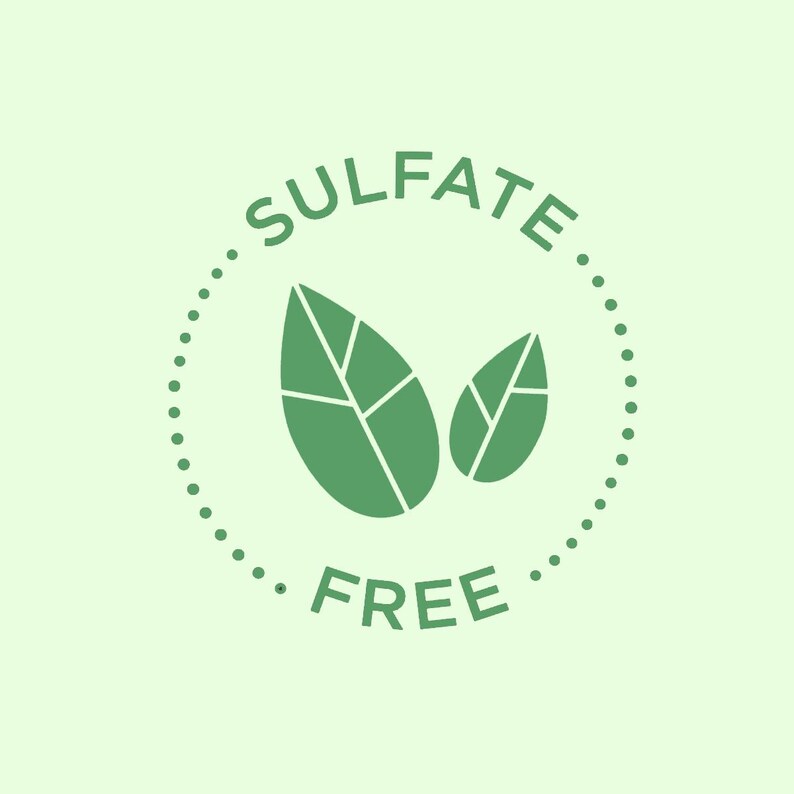 Op de afbeelding: Een groene cirkel met een witte gestippelde rand en de tekst "Sulfate Free" in wit. Twee groene bladeren bevinden zich in de cirkel.