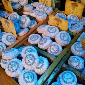May include: A variety of wrapped soaps in wooden crates. The soaps are white with blue and gold accents. The soaps are arranged in rows and are labeled with the text "Shampoo Bar".