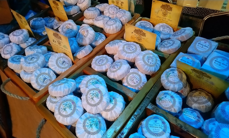 May include: A variety of wrapped soaps in wooden crates. The soaps are white with blue and gold accents. The soaps are arranged in rows and are labeled with the text "Shampoo Bar".
