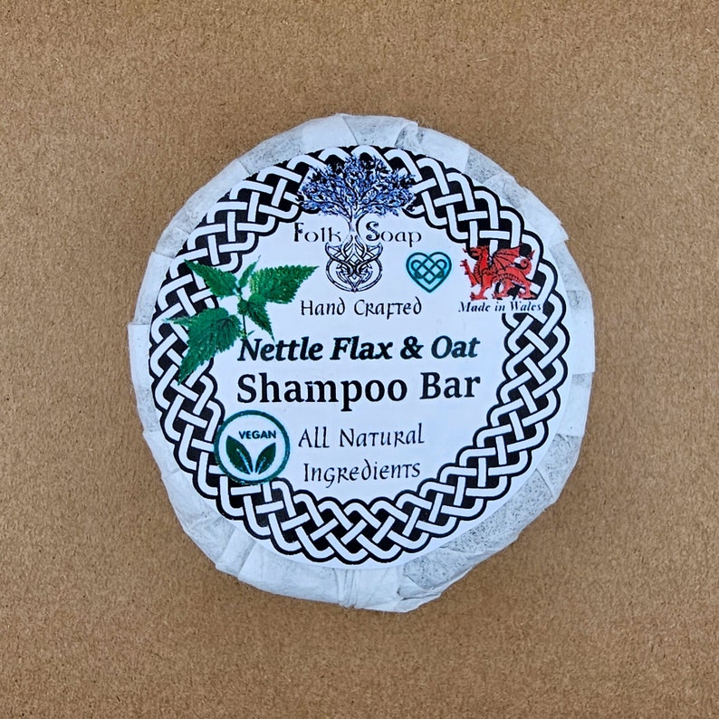 May include: A white round bar of soap with black Celtic knot border. The label reads "Folk Soap", "Hand Crafted", "Made in Wales", "Nettle Flax & Oat Shampoo Bar", "Vegan", "All Natural Ingredients".