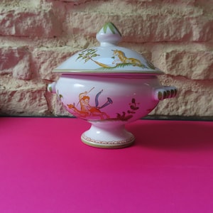 Op de afbeelding: Een roze keramische serveerschaal met deksel en voet. De schaal is versierd met een geschilderde scène van een figuur met pijl en boog en een mythisch wezen. Het deksel heeft een vergelijkbaar ontwerp en een groene knop.