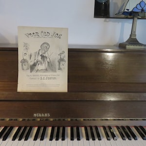 Peut inclure: Une partition musicale vintage pour la chanson "Poor Old Joe" avec une illustration en noir et blanc d'un homme tenant une bouteille et un bâton. La partition est posée sur un piano en bois brun avec un clavier noir et blanc.