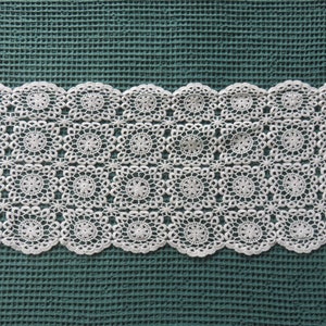 Peut inclure: Napperon en dentelle blanche crochetée avec un motif carré. Le napperon est sur une surface texturée verte.