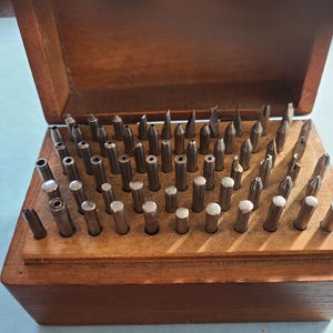 May include: A wooden box with a hinged lid, filled with an assortment of small, metal tools. The tools are various shapes and sizes, including pointed, flat, and cylindrical. The box is made of dark wood and is open, revealing the tools inside.
