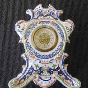 Pode incluir: Um relógio de cerâmica branca com desenhos florais azuis, verdes e laranja. O mostrador do relógio é branco com algarismos romanos pretos e ponteiros dourados.