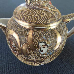 Op de afbeelding: Een antieke, sierlijke theepot met een deksel, tuit en handvat. De theepot is voornamelijk goud en bruin met ingewikkelde patronen en ontwerpen. Het heeft een geschilderd gezicht aan de zijkant en is waarschijnlijk gemaakt van keramiek of porselein.