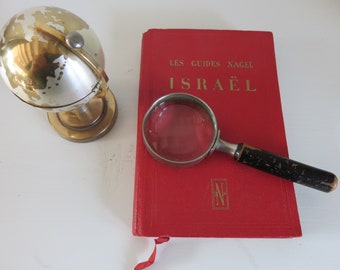May include: A red book titled "LES GUIDES NACEL ISRAEL" with a magnifying glass on top of it. A small gold globe is in the background.