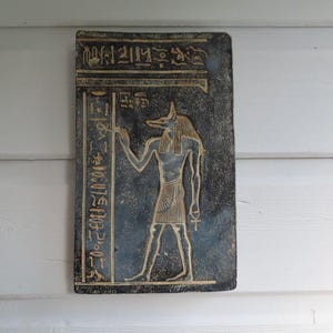 Könnte beinhalten: Eine rechteckige, dunkelblaue Tafel mit goldenen Hieroglyphen und einer Anubis-Figur. Die Anubis-Figur steht und hält einen Stab. Die Tafel ist wahrscheinlich ein Dekorationsartikel oder ein Wandbehang.