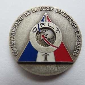 Może przedstawiać: Okrągły srebrny medalion z trójkątnym wzorem w kolorze niebieskim, białym i czerwonym. Medalion ma napis "COMMANDE DE LA FORCE LOGISTIQUE TERRESTRE" wokół krawędzi. W środku medalionu znajduje się okrąg z napisem "O.F.L.T." i globus ze strzałką skierowaną do góry.