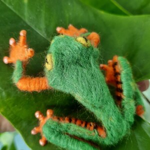 May include: A handmade felt frog with a bright green body, orange limbs, and yellow eyes rests on a large green leaf. The frog's feet and legs have black and orange stripes. The frog is a whimsical, handcrafted item.
