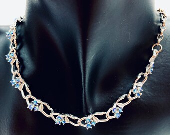 May include: Silver-toned necklace with a floral design. The necklace features small clusters of blue gemstones and leaf-shaped accents. The necklace is displayed on a black mannequin, showcasing its delicate details and elegant style.