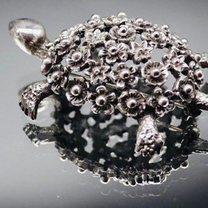 Puede incluir: Broche plateado con forma de tortuga. El caparazón está adornado con pequeños diseños en forma de flor. El broche tiene una superficie reflectante, creando un efecto de espejo. La cabeza y las patas de la tortuga son visibles, lo que aumenta su realismo.