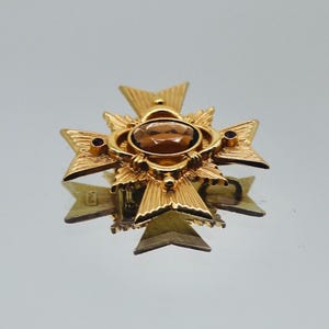 Pode incluir: Broche dourada em forma de cruz, com uma pedra oval âmbar no centro. A broche tem um padrão texturizado de estrela e pequenos detalhes em vermelho escuro. Um acessório de estilo vintage.