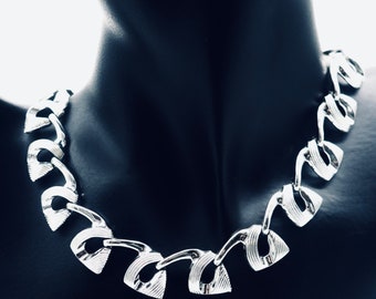 May include: A silver-toned necklace with a geometric design. The necklace features a series of interconnected, stylized links with a textured surface. The necklace is displayed on a black mannequin, highlighting its metallic sheen and intricate details.