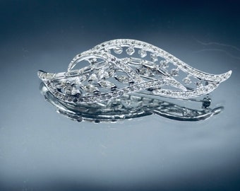 May include: A silver-toned brooch in a leaf design, encrusted with clear crystals. The brooch has a pin closure on the back and is displayed on a reflective surface. The crystals are set in a delicate, openwork pattern.
