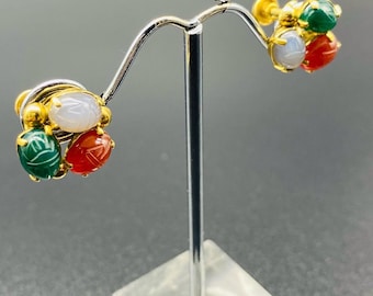 Könnte beinhalten: Goldfarbene Ohrringe mit drei farbigen, geschnitzten Steinakzenten: grün, rot und weiß. Die Ohrringe haben einen Steckerverschluss und werden auf einem silbernen Ständer vor grauem Hintergrund präsentiert.