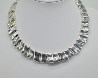 May include: A silver necklace featuring a series of textured, rectangular links. The links are arranged in a curved shape, creating a bold, modern design. The necklace is displayed on a white surface, highlighting its metallic sheen.