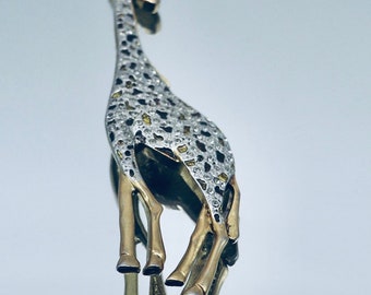 Könnte beinhalten: Eine giraffenförmige Brosche mit goldfarbenem Metallfinish an Beinen und Kopf sowie einem strukturierten Körper, der mit kleinen, funkelnden klaren Kristallen und dunklen, unregelmäßigen Flecken besetzt ist, die das Fellmuster nachahmen.