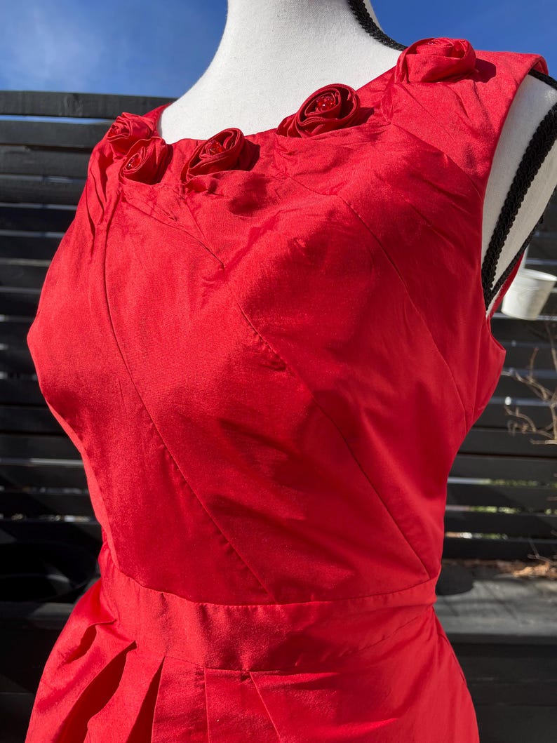 Puede incluir: Un vestido rojo sin mangas con un corpi&ntilde;o ajustado y una falda completa. El escote est&aacute; adornado con cinco grandes rosas de tela roja.