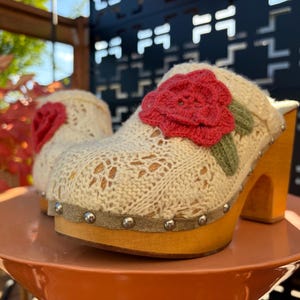 Op de afbeelding: Een paar crèmekleurige gebreide klompen met een opvallende rode gehaakte roosapplicatie met groene bladeren. De schoenen hebben een lichtgekleurde houten plateauzool en een beige suède rand die is vastgezet met zilverkleurige metalen studs.