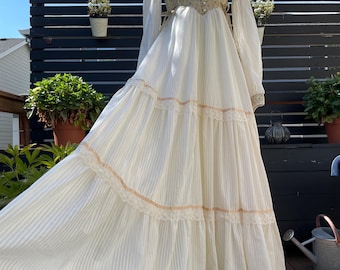 May include: A vintage-style cream-colored dress with vertical stripes and lace trim. The bodice features a floral pattern and button details. Long, sheer sleeves and a flowing skirt create a romantic silhouette. The dress is displayed on a mannequin outdoors.