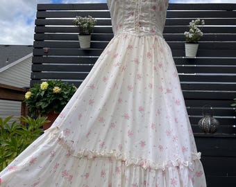 Puede incluir: Un vestido blanco de estilo vintage adornado con un delicado estampado de rosas rosas. El vestido presenta un corpiño con ribete de encaje, un frente estilo corsé y una falda fluida con volantes. El vestido se exhibe en un maniquí.