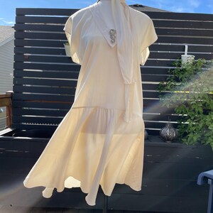May include: A white, sleeveless dress with a ruffled hem and a tie at the neck. The dress has a decorative brooch on the front.