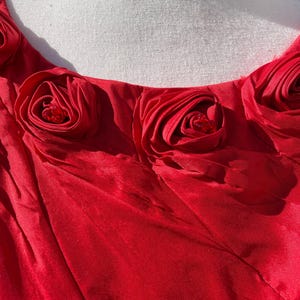 Puede incluir: Vestido rojo con escote redondo y una fila de rosas de tela roja con centros de pedrer&iacute;a.