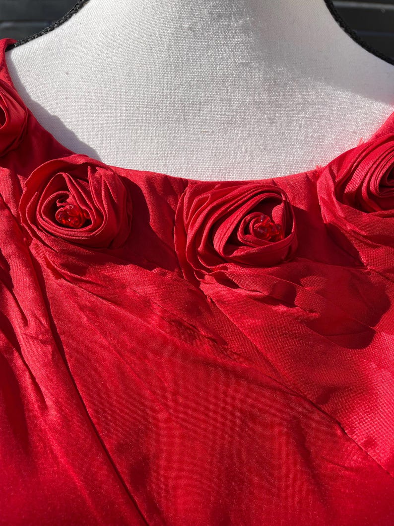 Puede incluir: Vestido rojo con escote redondo y una fila de rosas de tela roja con centros de pedrer&iacute;a.