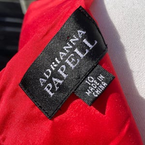 Puede incluir: Una etiqueta negra con texto blanco que dice "Adrianna Papell" y "Hecho en China" sobre un fondo de tela roja.