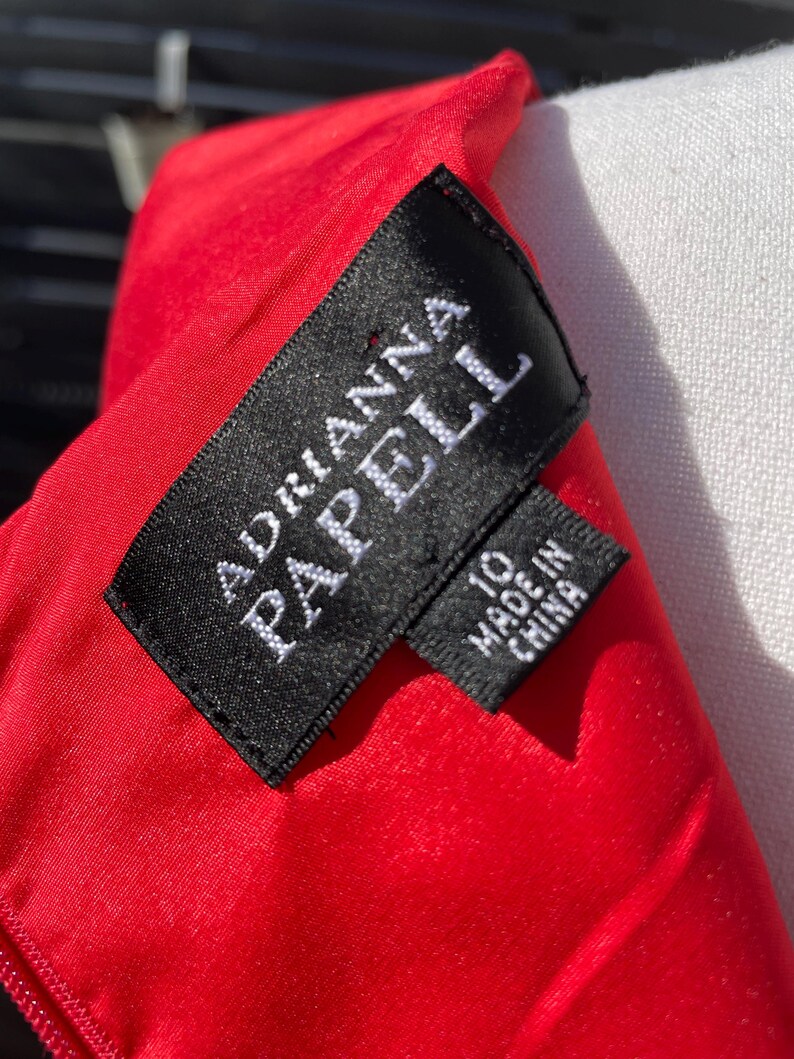 Puede incluir: Una etiqueta negra con texto blanco que dice "Adrianna Papell" y "Hecho en China" sobre un fondo de tela roja.
