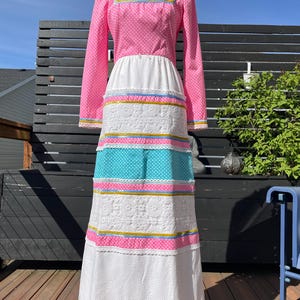 Puede incluir: Un vestido largo rosa y blanco con una franja azul y blanca y ribete de encaje. El vestido tiene un escote cuadrado y mangas largas.