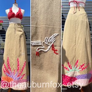 以下が含まれることがあります： 白い刺繍の鳥のデザインとピンク、パープル、グリーンの生地の継ぎ接ぎの裾を持つ、長いベージュのスカート。スカートはハイウエストでストレートシルエットです。