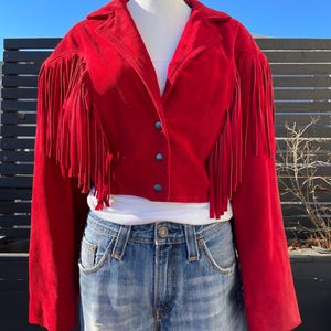 May include: A red suede jacket with fringe detailing on the sleeves and hem. The jacket is cropped and has a button closure.