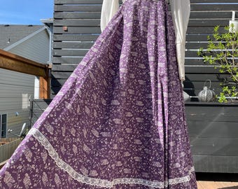 May include: A long, purple floral print dress with white lace trim and long white sleeves. The dress has a scoop neckline and a fitted bodice. The skirt is full and flowing.