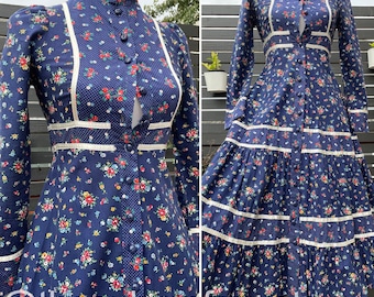 Puede incluir: Un vestido largo de manga larga, de flores azules, con ribetes de encaje blanco y un frente abotonado. El vestido tiene un corpiño ajustado y una falda fluida con múltiples niveles.