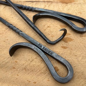 May include: Three handcrafted, dark metal fire pit tools. Two have curved handles and the text "Josh" and "Mr Brown" engraved on them. One tool has a twisted handle design.